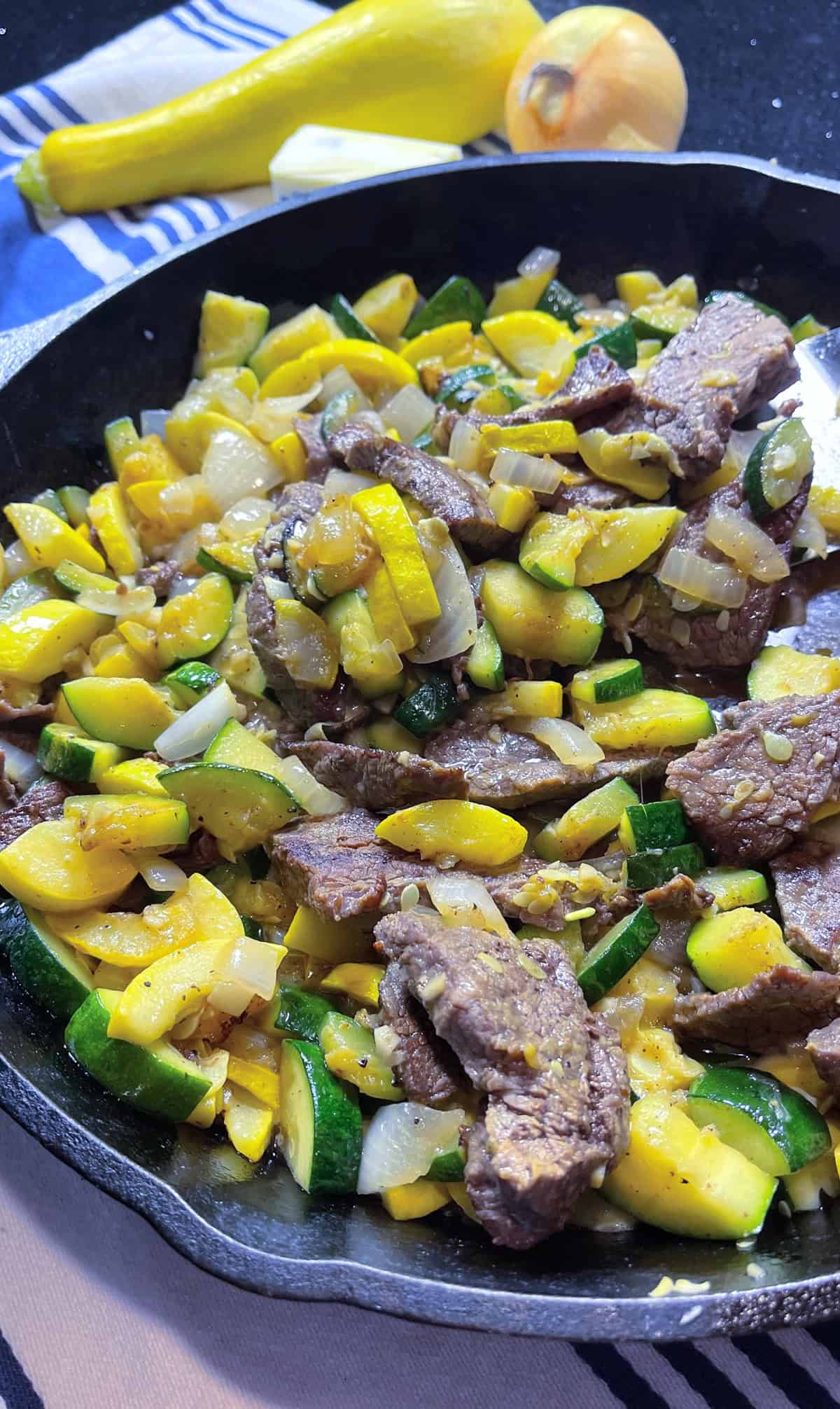 Browned flank steak slices in a cast iron skillet with onions, zucchini, and summer squash.