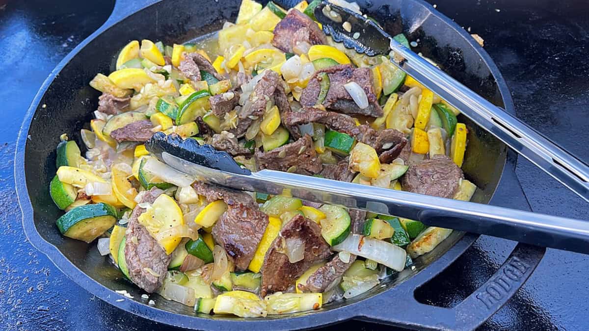 A cast iron skillet on a griddle filled with cooked flank steak and squash.