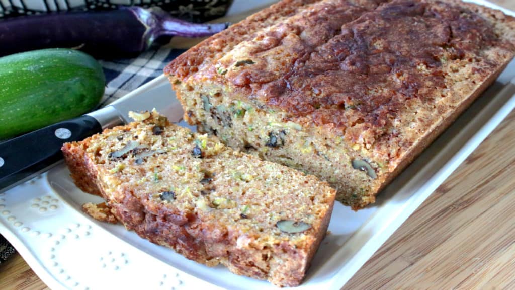 A loaf of sliced eggplant zucchini bread on a white rectangular plate with a eggplant and zucchini in the background.