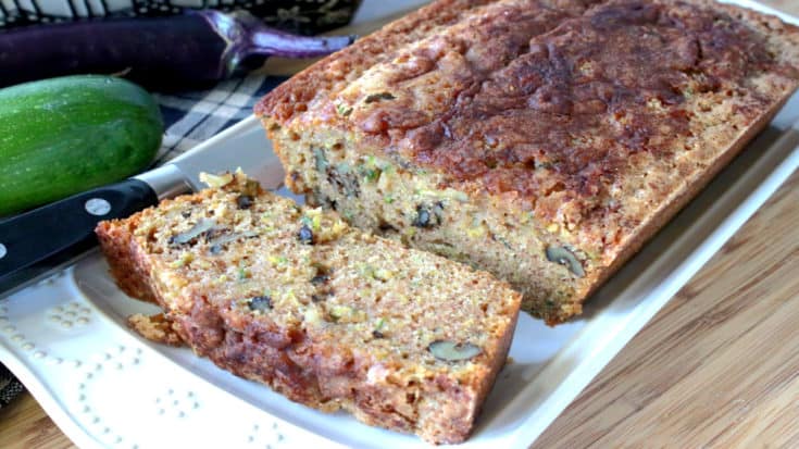 A loaf of sliced eggplant zucchini bread on a white rectangular plate with a eggplant and zucchini in the background.