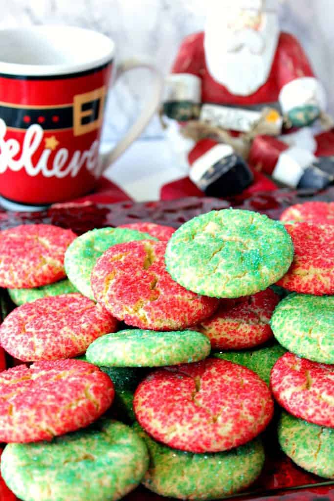 A vertical closeup photo of red and green sugar cookies with Santa and a coffee mug in the background.