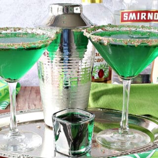 A couple of martini glasses on a silver tray filled with green Irish Grasshopper Cocktails along with a cocktail shaker and a shot glass.