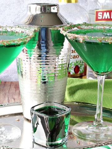 A couple of martini glasses on a silver tray filled with green Irish Grasshopper Cocktails along with a cocktail shaker and a shot glass.