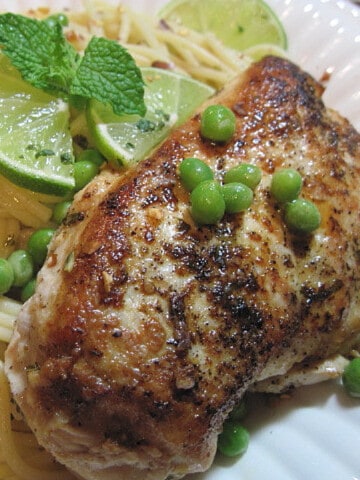 A Mojito Chicken breast on a white plate with lime slices, mint, peas, and spaghetti.