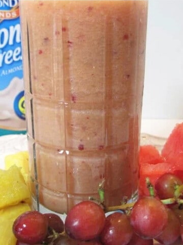A Fruit Smoothie in a tall glass with pineapple, grapes, and watermelon surrounding it.
