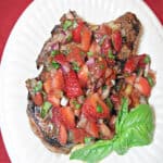 An overhead photo of a Pork Chop with Strawberry Relish on a white plate with fresh basil as garnish.