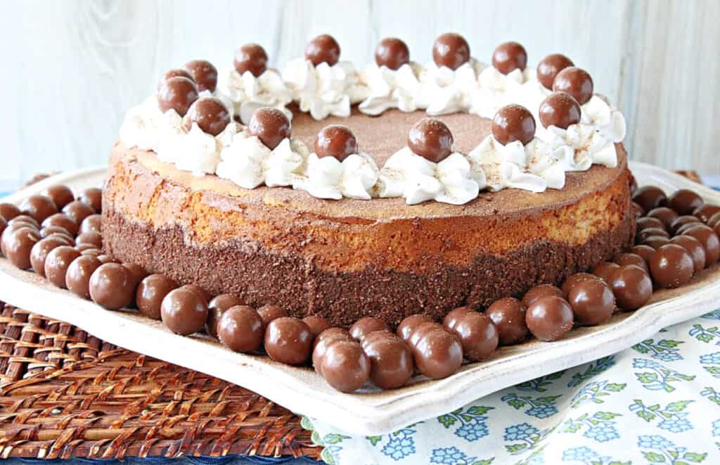 An entire Malted Milk Ball Cheesecake on a square white plate surrounded by malted milk balls.