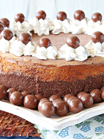 An entire Malted Milk Ball Cheesecake on a square white plate surrounded by malted milk balls.