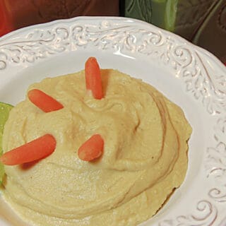 A pretty white bowl filled with creamy White Bean and Artichoke Hummus with baby carrots and a lime wedge.