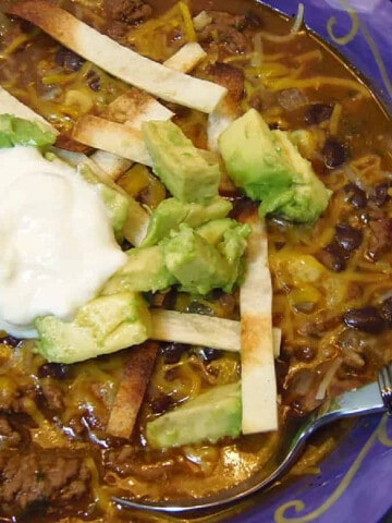 A purple bowl filled with a serving of Black Bean Soup and topped with avocado and sour cream.