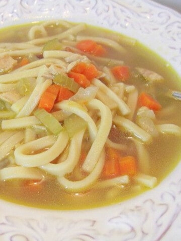 A pretty white bowl filled with Homemade Chicken Noodle soup with carrots and celery.