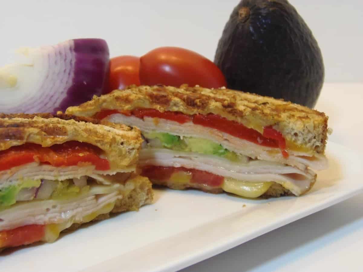 A colorful layered Turkey and Chicken Panini Sandwich with tomatoes, avocado, cheese, and meat.