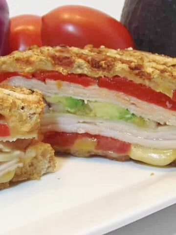 A colorful layered Turkey and Chicken Panini Sandwich with tomatoes, avocado, cheese, and meat.