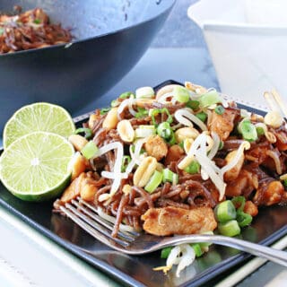 A serving of Chicken Pad Thai on a black plate with a lime, fork, and chopsticks.