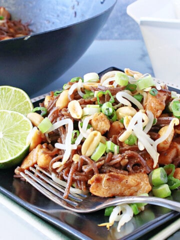 A serving of Chicken Pad Thai on a black plate with a lime, fork, and chopsticks.