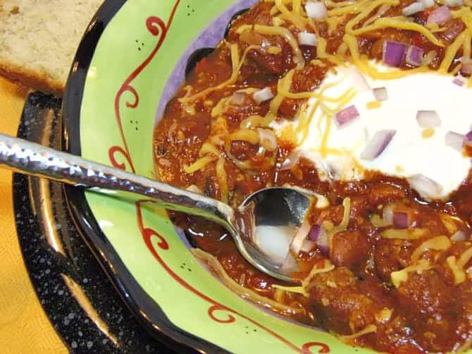 Closeup of Smoky Beer Chili in a bowl with a spoon with cheese and onions - kudoskitchenbyrenee.com