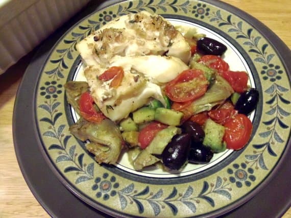A serving of Mediterranean Baked Cod with artichokes, olives, and tomatoes on a dinner plate.