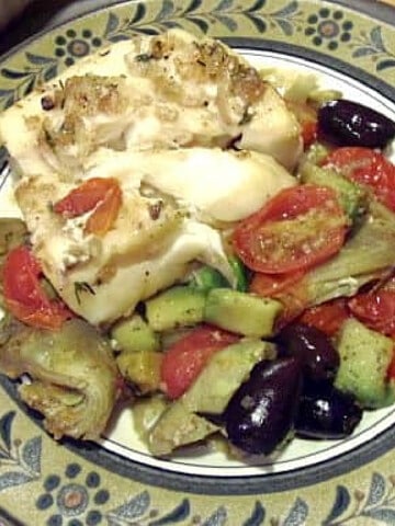 A serving of Mediterranean Baked Cod with artichokes, olives, and tomatoes on a dinner plate.