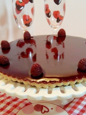 A shiny Raspberry Custard Tart on a cake plate with champagne flutes in the background.