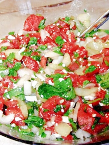 A glass bowl filled with Watermelon Cucumber Salad along with serving tongs.