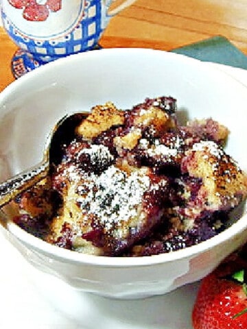 A white bowl filled with Blueberry Grunt along with strawberries on the side and a spoon in the bowl.