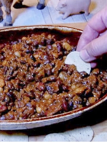 An oval brown bowl filled with Barn Dance Bean Dip and a hand holding a chip.