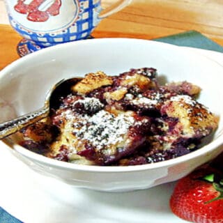 A white bowl filled with Blueberry Grunt along with strawberries on the side and a spoon in the bowl.