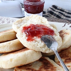A platter of Homemade English Crumpets and one with strawberry jam and a spoon.