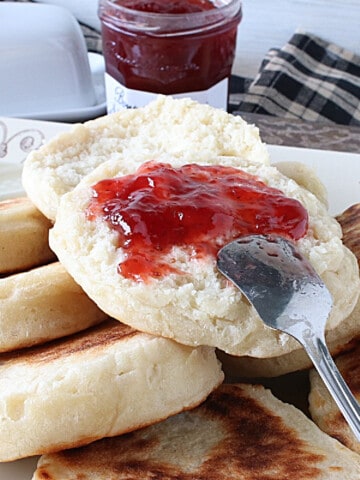 A platter of Homemade English Crumpets and one with strawberry jam and a spoon.