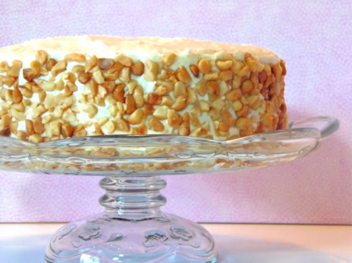 A whole banana poppy seed cake on a glass cake stand covered in chopped macadamia nut cookies