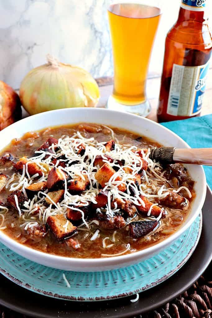 A bowl of German onion soup with cheese, bratwurst, pretzel croutons, and a glass of beer in the background.