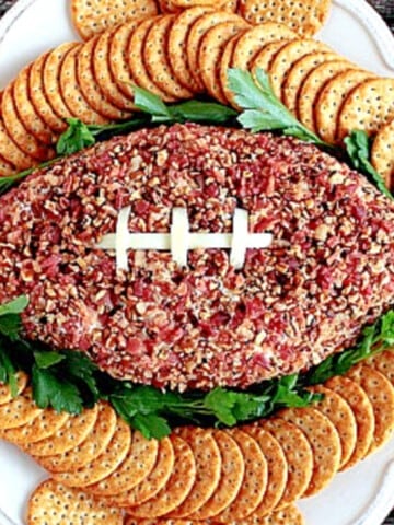 An overhead photo of a Football-Shaped Cheeseball with Pecans, crackers, and parsley on a platter.