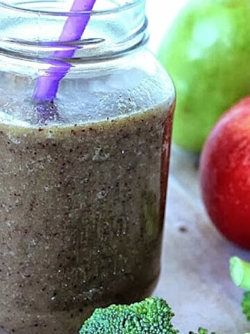 A Fruit and Veggie Smoothie in a mason jar with a purple straw and broccoli and fruit on the side.