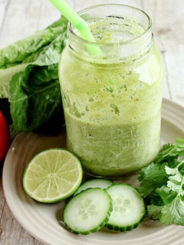 A Healthy Green Smoothie in a mason jar on a plate with lime, cucumber, and parsley.