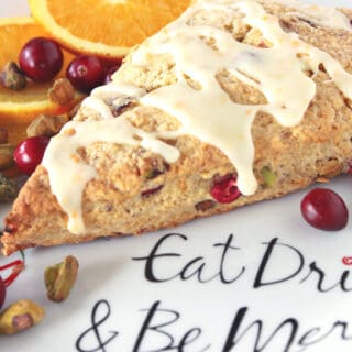 A vertical closeup of a Cranberry Orange Scone with Pistachios along with a glaze on a festive plate.
