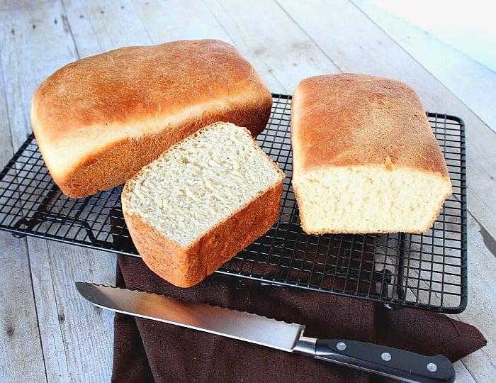 A horizontal photo of two loaves of buttermilk honey bread on a cooling rack with a serrated knife.