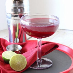 A Mixed Berry Margarita on a red serving tray with a lime and cocktail shaker.