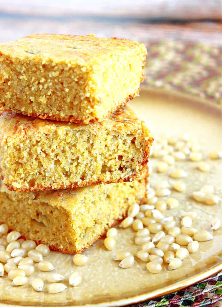 A vertical photo of a stack of three pieces of Buttermilk Green Chile Cornbread on a plate along with some kernels of corn.