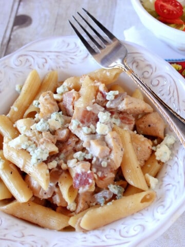 A white bowl filled with Chicken Penne Pasta and topped with bacon and blue cheese.
