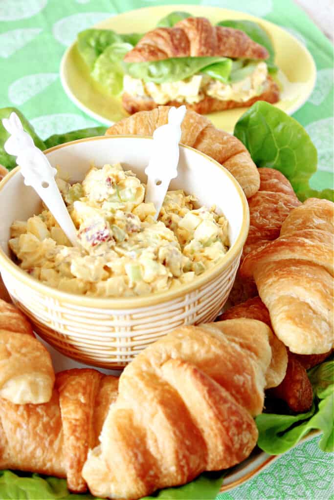 A small bowl with two bunny spoons filled with creamy Bacon Egg Salad surrounded by baked croissants.