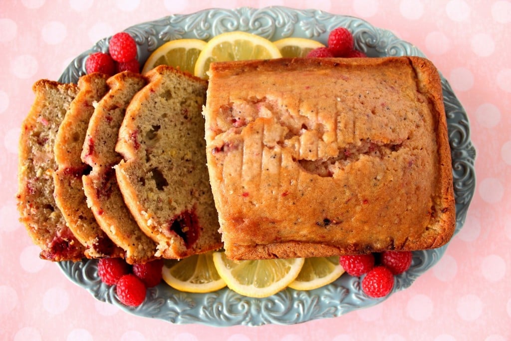 A direct overhead photo of a loaf of sliced lemon raspberry bread on a blue platter with lemon slices and fresh raspberries on the platter.