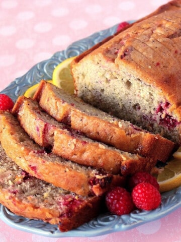 A Lemon Raspberry Cardamom Quick Bread on a blue plate with raspberries and lemon slices surrounding it.