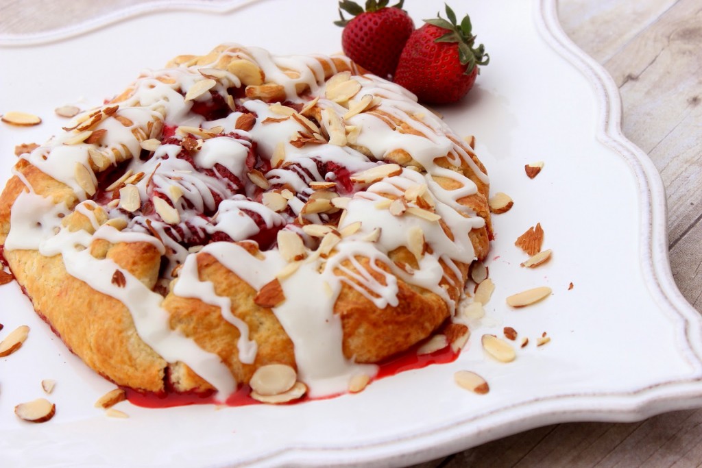Strawberry Crostata with Sweet Almond Glaze A horizontal photo of a strawberry crostata on a pretty white serving platter with fresh strawberries as garnish, and an almond glaze and slivered almonds