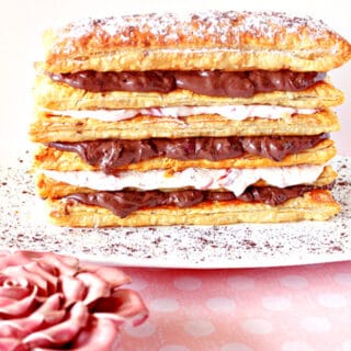 A Chocolate Cherry Pastry Tower on a white plate with a dusting of cocoa powder and a pink rose in the foreground..