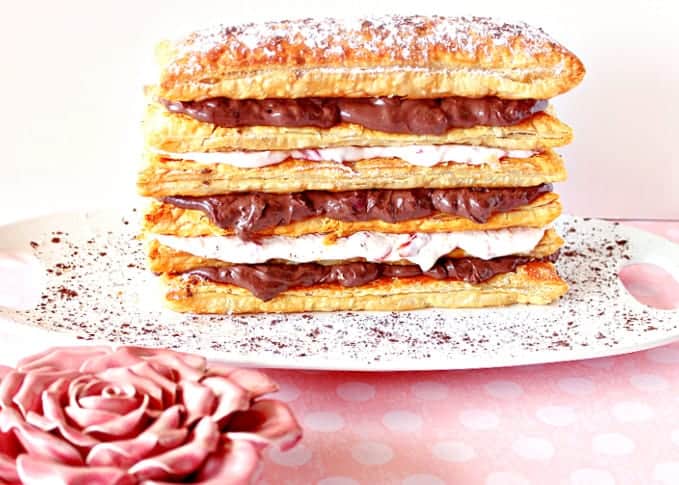 A Chocolate Cherry Pastry Tower on a white plate with a dusting of cocoa powder and a pink rose in the foreground..