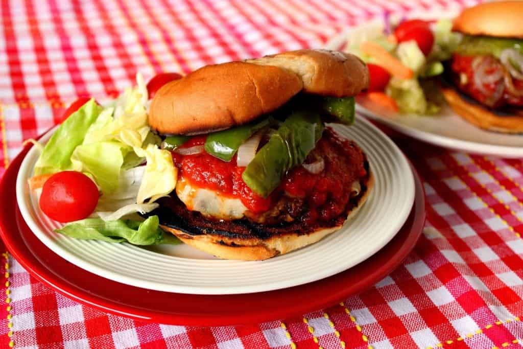 An Italian sausage pizza burger on a white plate with green peppers, lettuce, and tomatoes.
