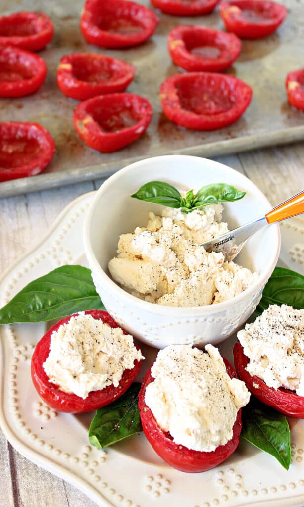 A tray of Roasted Roma Tomatoes in the background and a plate of Roasted Roma Tomatoes with Homemade Red Pepper Ricotta in the foreground with black pepper and basil.