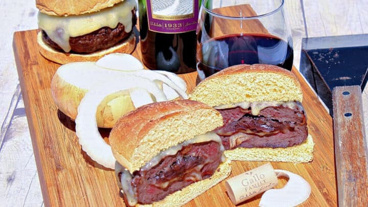 A sliced in half Burger Stuffed with Caramelized Onions on a cutting board along with a glass of wine and some onions.