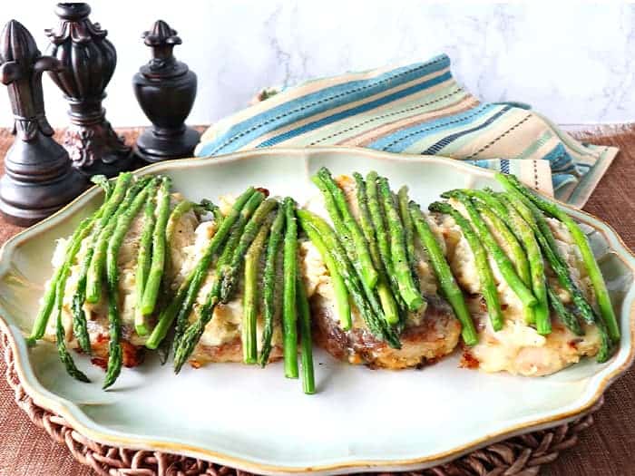 A horizontal photo of Chicken Oscar on a platter with asparagus spears and a blue and tan striped napkin. Valentine's day dinner recipe roundup.