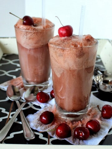 Two tall glasses of Chocolate Cherry Cows on a tray with straws and cherries.